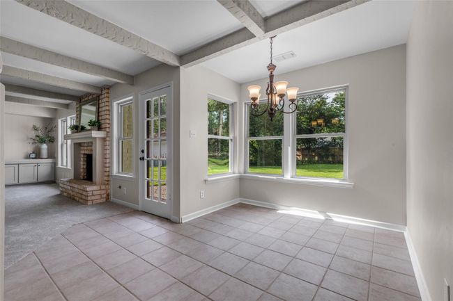 Bright breakfast area with large windows and a view of the yard, featuring tiled floors and a stylish chandelier. Adjacent to a cozy living space with a brick fireplace and built-in shelving. | Image 22