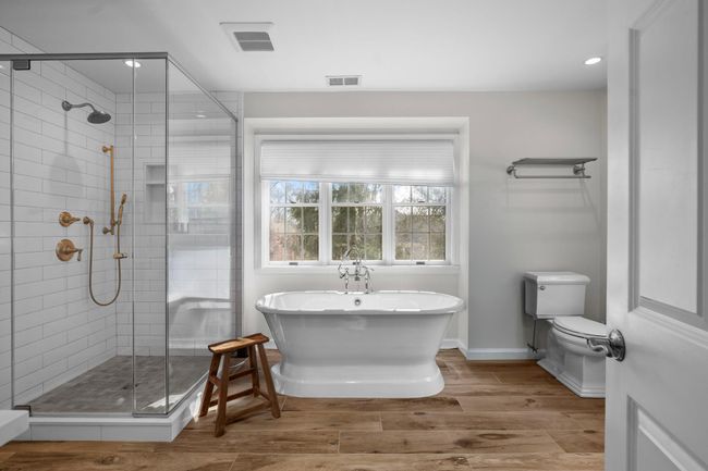 The spa-like primary en suite features a freestanding soaking tub positioned beneath a picture window, complemented by a sleek glass-enclosed shower with elegant brass fixtures. | Image 24