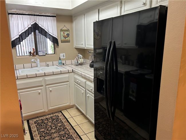 Kitchen with black refrigerator with ice dispenser, tile countertops, white cabinetry, and light tile patterned floors | Image 13