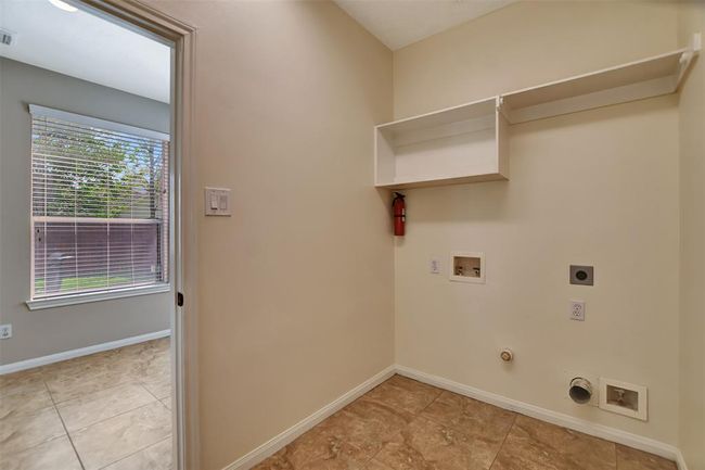 This photo shows a laundry room with hookups for a washer and dryer, wall shelving for storage, and tile flooring. A window in the adjoining room lets in natural light, and a fire extinguisher is mounted on the wall. | Image 27