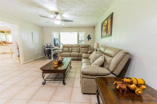 This inviting living room features tiled floors, and a ceiling fan. There's ample natural light from a large window, and it connects seamlessly to the kitchen area. Modern decor and neutral tones create a cozy atmosphere. | Image 11