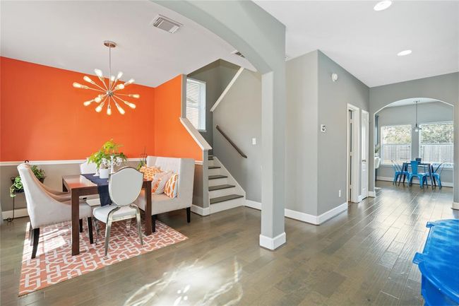 This photo showcases a modern dining area with a vibrant orange accent wall and stylish light fixture. It features a cozy dining set with a bench, adjacent to a staircase with neutral gray walls. The space leads to another room with large windows and ample natural light, creating a warm and inviting atmosphere. | Image 24