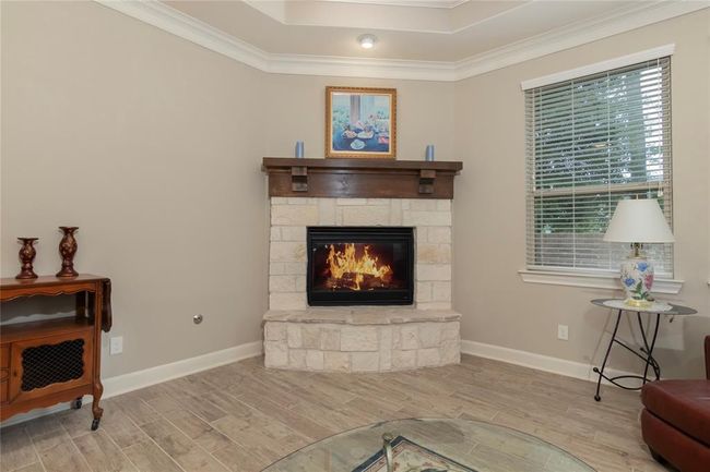 Cozy living room corner featuring a stone fireplace with a wooden mantel, a large window with blinds providing natural light, and elegant crown molding. The room has a warm, inviting atmosphere with neutral walls and wood-look flooring. | Image 11