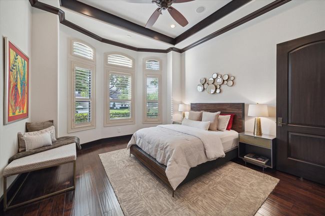 This sunny secondary bedroom enjoys a curved wall of windows with plantation shutters, allowing abundant natural light while maintaining privacy. The clean, modern aesthetic is enhanced by a trayed ceiling with dark wood trim, rich hardwood flooring, and a warm neutral palette. Featuring an ensuite bathroom, walk-in closet with built-ins, and plush textures, this space feels inviting, yet functional—perfect for a guest or family member to unwind in comfort. | Image 34