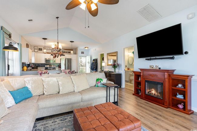 Living area featuring light wood finished floors, visible vents, vaulted ceiling, ceiling fan with notable chandelier, and a glass covered fireplace | Image 6