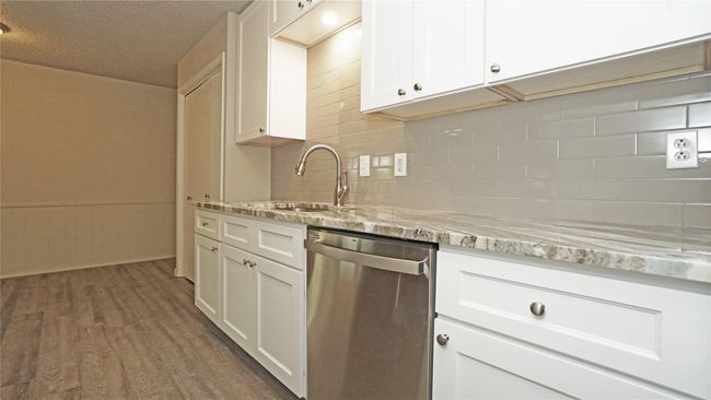 The kitchen with a stainless steel dishwasher, gorgeous granite counters, tile backsplash, white cabinetry, and luxury vinyl plank floors opens to the dining area | Image 8