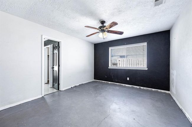 This room features a minimalist design with concrete flooring and a two-tone wall, accentuated by a black accent wall. It includes a ceiling fan and a window with blinds for natural light. The space is adjacent to a bathroom, accessed through a door with a dark finish. | Image 17