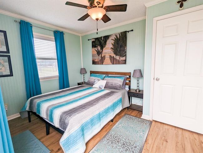 Bedroom with a beach-themed decor, featuring a double bed with a striped comforter and seating futon. There's a ceiling fan and ample natural light from a window. A beach artwork above the bed adds a relaxing touch. | Image 13