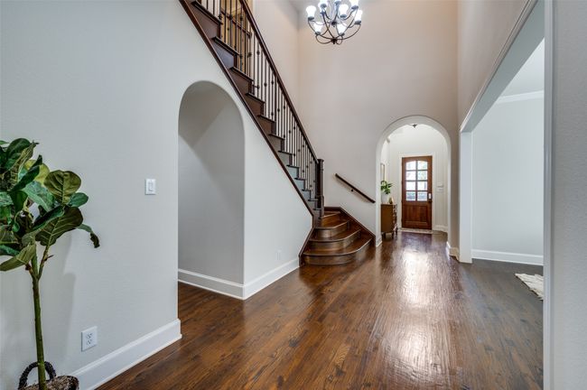 The foyer features a soaring two-story ceiling, arched walkways, and dark wood-finished floors with elegant baseboards. A striking chandelier illuminates the space, while the beautiful staircase with wrought iron railing adds a touch of grandeur. | Image 6