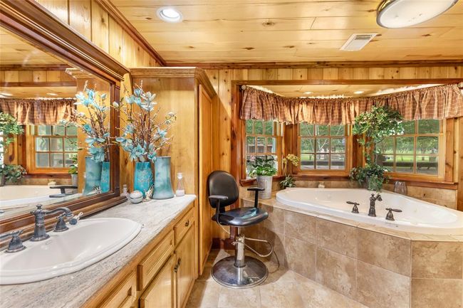 This rustic bathroom features a cozy wooden interior with a large, inviting corner bathtub surrounded by windows. It includes a stylish vanity with twin sinks and decorative mirrors. The natural light and greenery create a serene atmosphere. | Image 32
