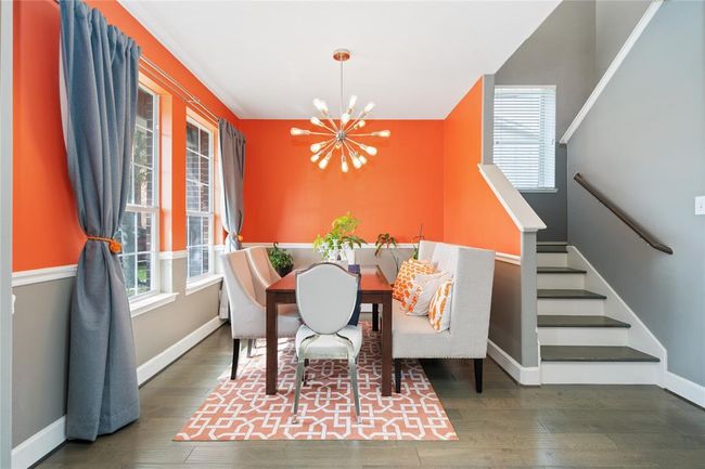 This vibrant dining area features bright orange walls and stylish gray accents, complemented by large windows with gray curtains. It includes a modern light fixture, a cozy dining setup with a cushioned bench, and an accent chair. The space is adjacent to a staircase, offering a contemporary and inviting atmosphere. | Image 10