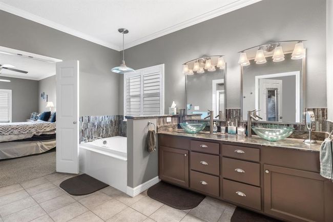 This photo showcases a modern bathroom with a dual sink vanity featuring glass vessel sinks, ample cabinet space, and elegant lighting. There's a tiled floor and a cozy bathtub area with decorative accent tiles. The room has a neutral color scheme and an adjoining view into a bedroom, enhancing the spacious, open feel. | Image 19