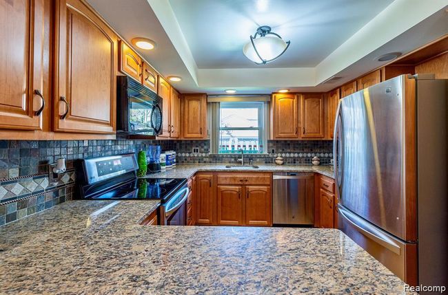 Kitchen with brown cabinetry, stainless steel appliances, light stone counters, a tray ceiling, and backsplash | Image 4