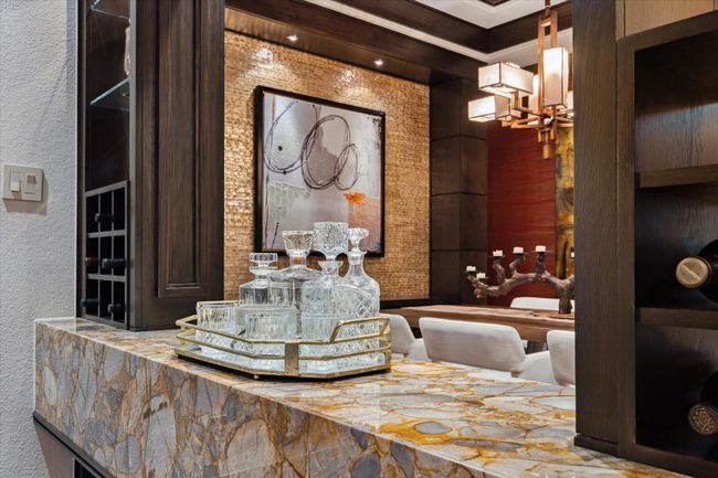 This sophisticated serving bar, seamlessly positioned between the family room and dining area, is a true statement piece. A richly veined imported onyx waterfall countertop anchors the space with luxurious texture and eye-catching depth. Open shelving and built-in wine storage blend form and function, while a backdrop of shimmering tile and bold artwork creates a cohesive visual connection to the adjoining dining room. Designed for effortless entertaining with a hint of glamour. | Image 11