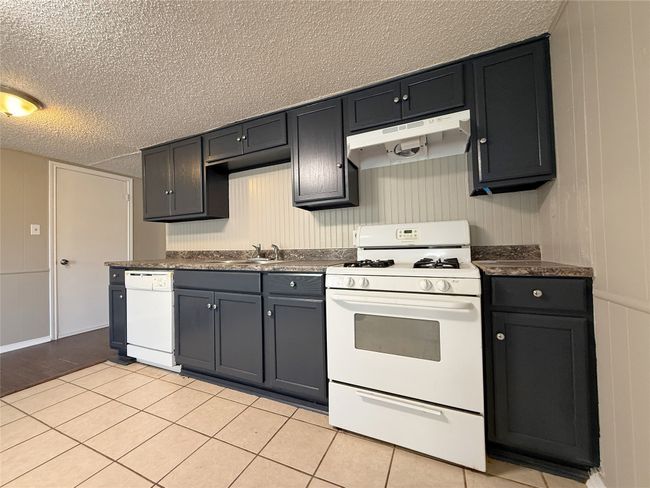 Kitchen with a textured ceiling, white appliances, dark countertops, under cabinet range hood, and a sink | Image 6