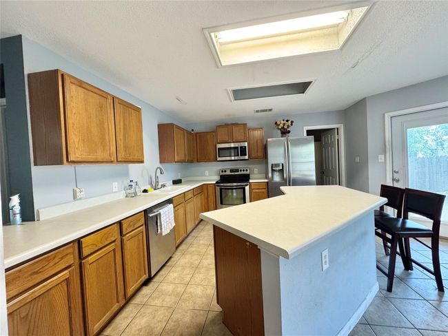 Kitchen with light tile patterned flooring, appliances with stainless steel finishes, brown cabinetry, a center island, and a textured ceiling | Image 5