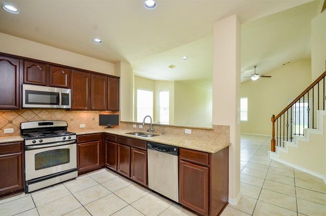 This photo showcases a modern kitchen with stainless steel appliances, dark wood cabinetry, and a tiled backsplash. The open layout connects to a bright living area. | Image 12