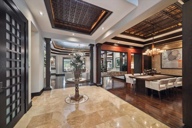 Step through the elegant entryway into this captivating transition space where layered coffered ceilings with intricate custom millwork and ambient lighting create architectural flair overhead, which truly emulates the finest of five-star resorts. Flanked by stately columns and 8-foot custom wood doors with milk glass panes, the open layout effortlessly connects the formal dining room and living areas, all unified by a blend of polished travertine and wide-plank hardwood flooring. | Image 8