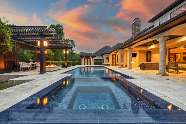 This breathtaking backyard retreat is anchored by a gleaming pool with a deep blue PebbleTec surface and raised infinity spa, accented with deep blue mosaic tile and surrounded by a sprawling travertine patio. At one end, a striking pergola frames a cozy fire pit lounge—perfect for sunset gatherings. Elegant stone columns support the expansive covered patio, complete with an outdoor fireplace and multiple seating areas. Ambient lighting and lush landscaping complete the resort-like ambiance of this private | Image 42