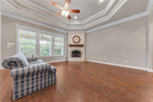 Living room featuring a tray ceiling, wood finished floors, a ceiling fan, a stone fireplace, and baseboards | Image 5