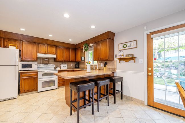 Kitchen featuring backsplash, light countertops, a peninsula, under cabinet range hood, and white appliances | Image 19