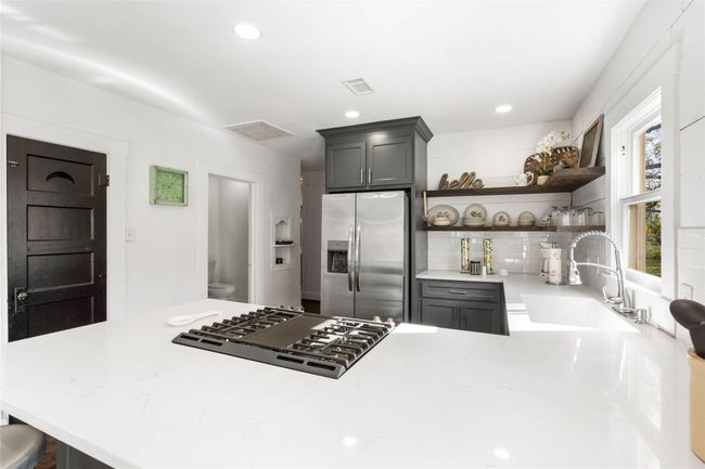 Kitchen featuring stainless steel fridge with ice dispenser, gas cooktop, open shelves, backsplash, and light stone counters | Image 13