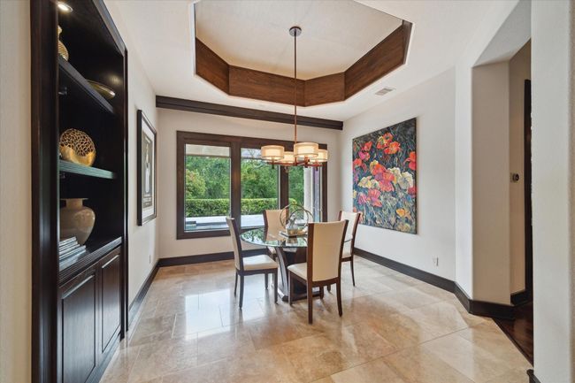 Flooded with natural light, this inviting breakfast room features a deep octagonal tray ceiling trimmed with warm wood accents, designer drum lighting, and polished travertine staggered flooring. A trio of picture windows frames peaceful views of the backyard and pool area, creating the perfect setting for morning coffee or casual meals. A built-in rich wood display cabinet adds elegance and functional storage, while colorful art brings the space to life. | Image 20
