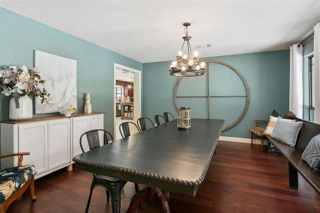 This dining room features a long, elegant table with mixed seating, accentuated by a modern chandelier. The teal walls and large circular wall decor add a stylish touch, complemented by hardwood flooring and a sideboard with decorative pieces. | Image 15