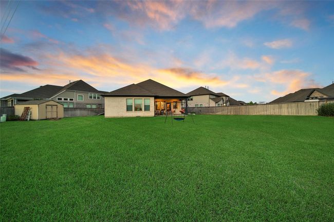 This photo showcases a spacious backyard with lush green grass and a swing set, perfect for outdoor activities. The single-story home features a cozy patio and large windows, set against a beautiful sunset sky. | Image 42