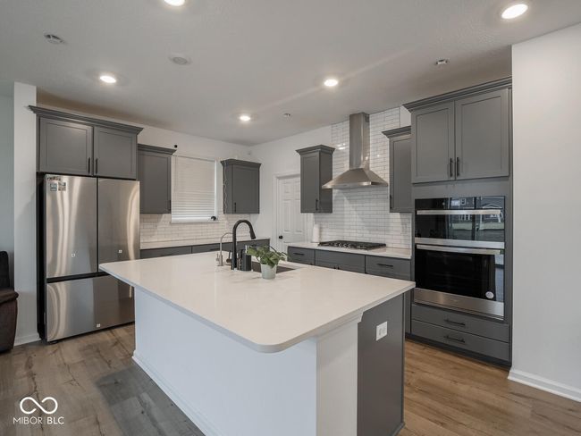 kitchen with wall chimney range hood, a center island with sink, stainless steel appliances, a sink, and wood finished floors | Image 18