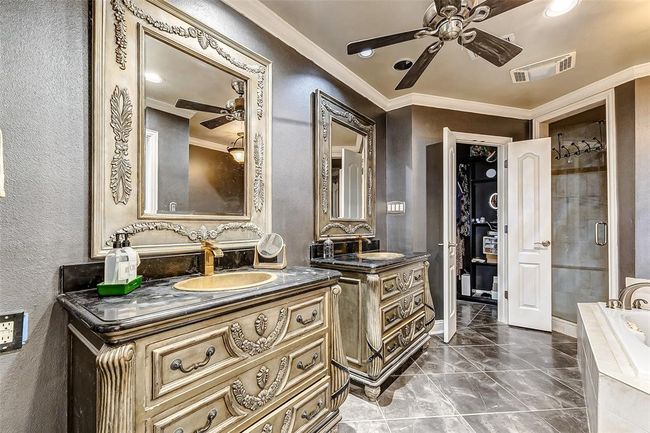 Luxury meets functionality in this beautifully appointed bathroom. Twin vanities with ornate detailing sit side by side, each with an oversized mirror and ample under-storage. A ceiling fan ensures comfort, while a jetted tub and an oversized shower with dual showerheads provide a spa-like experience. Soft gray walls complement the tile flooring, creating a serene and refined atmosphere. | Image 27