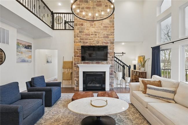 Stunning two-story great room featuring a floor-to-ceiling stone fireplace, dramatic wall of windows with woodland views, and a grand chandelier. Open and airy layout with hardwood floors and abundant natural light, creating a warm and inviting space ideal for relaxing or entertaining. | Image 10