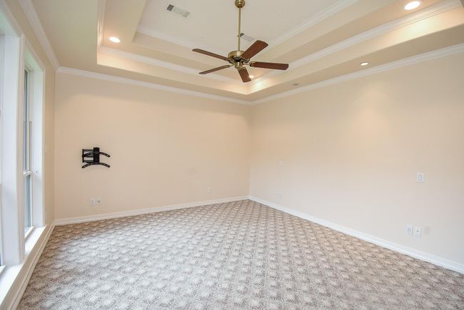 This room features a spacious design with a tray ceiling, recessed lighting, and a ceiling fan. It has neutral-colored walls and patterned carpet flooring, with large windows that allow natural light to brighten the space. A wall mount is available for a TV. | Image 33