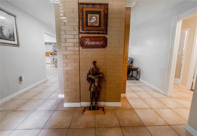 This photo shows a tiled foyer area with light walls and a central brick pillar. Open doorways lead to a kitchen and a living space, creating a welcoming and connected layout. | Image 6