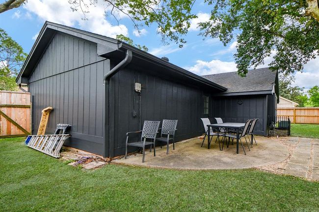 This photo shows a modern, dark-painted home exterior with a small patio area featuring outdoor seating. The yard is fenced and surrounded by greenery, providing privacy and a cozy outdoor space. | Image 43