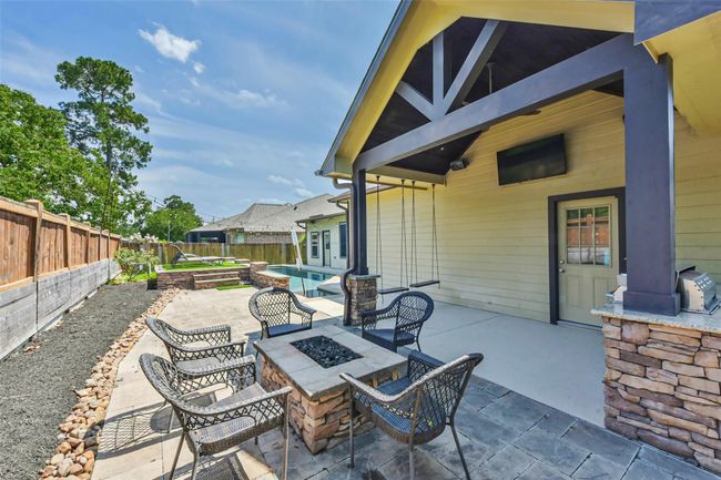 The outdoor space features a covered patio with a ceiling fan and mounted TV, a stone fire pit surrounded by seating, and a built-in grill. The area is paved, with a view of a landscaped garden and pool, offering a perfect setting for relaxation and entertainment. | Image 32