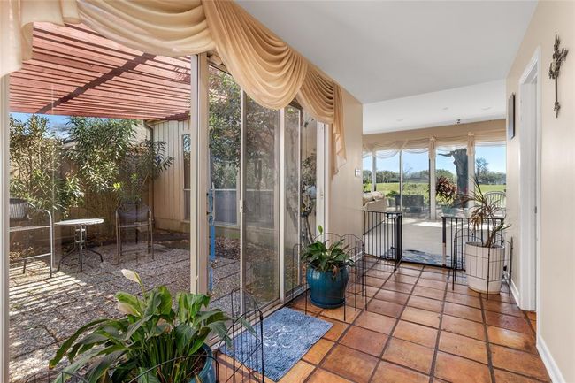 This bright and airy hallway features large sliding glass doors that open to a private center courtyard, allowing natural light to flow into the space. The courtyard offers a peaceful outdoor retreat with a paved patio and lush greenery. Further down, the hallway leads to a living area with expansive windows showcasing scenic views. | Image 9
