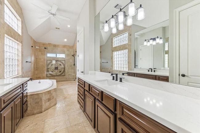 This spacious bathroom features a luxurious design with a large soaking tub, dual sinks with elegant fixtures, ample cabinetry, and a separate shower. Natural light filters through glass block windows, enhancing the bright and airy atmosphere. | Image 16