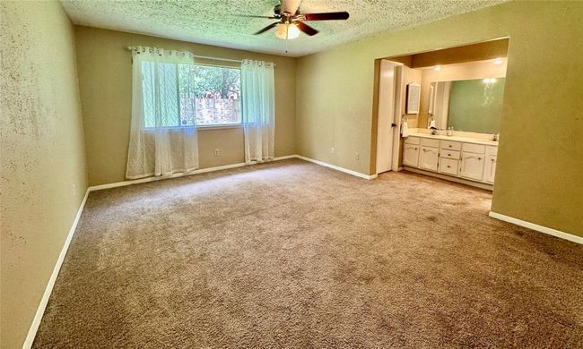 This spacious bedroom features carpet flooring, a ceiling fan, and ample natural light from a large window with sheer curtains. It includes an adjacent vanity area with a mirror and cabinetry, creating a convenient dressing space. | Image 10