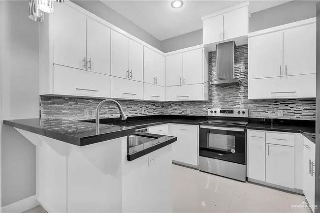 Kitchen with stainless steel range with electric cooktop, wall chimney range hood, dark countertops, a peninsula, and white cabinets | Image 9