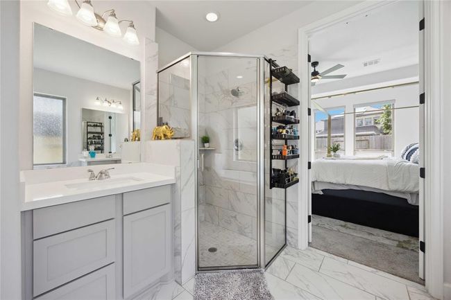 This photo showcases a modern bathroom with a glass-enclosed corner shower and elegant marble tile. A sleek vanity with ample storage and a large mirror is featured alongside stylish light fixtures. The bathroom connects to a bright bedroom with large windows, offering a cohesive and contemporary design. | Image 26
