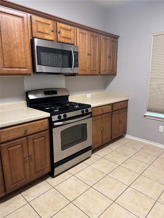 Kitchen with stainless steel appliances, brown cabinets, light countertops, light tile patterned floors, and baseboards | Image 4