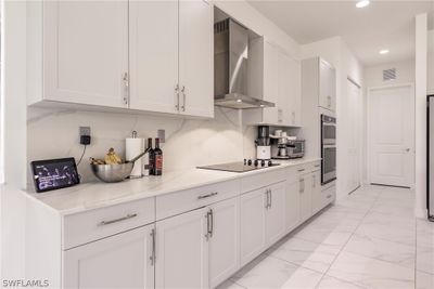 Kitchen with white cabinetry, wall chimney range hood, light stone countertops, black electric stovetop, and double oven | Image 2