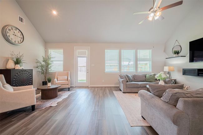 Bright and spacious living room with high ceilings. Features include wood-style flooring, a ceiling fan, and large windows that let in natural light. The room opens to an outdoor space through a glass door. | Image 11