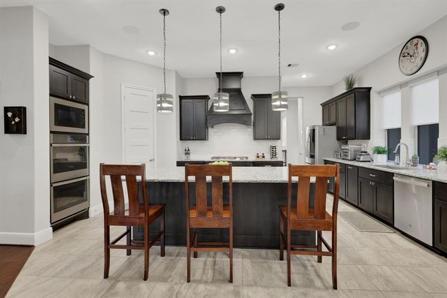 This modern kitchen features sleek dark cabinetry, a large granite island with seating, stainless steel appliances, and stylish pendant lighting. The space is bright and welcoming, ideal for cooking and entertaining. | Image 19