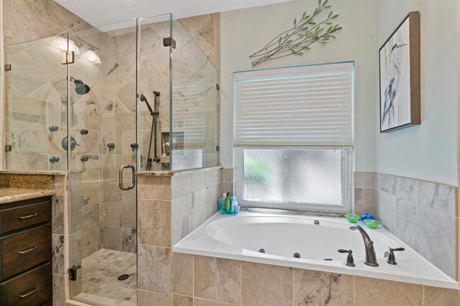 This luxurious primary bathroom features a spa-like design with elegant tile throughout, showcasing a spacious glass-enclosed shower with multiple body jets, separate handheld shower and rainhead, and a relaxing soaking tub positioned beneath a frosted window with natural light filtering through. | Image 20