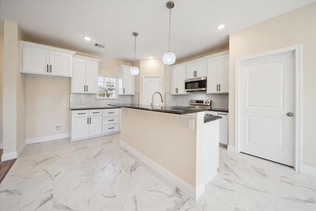 Kitchen featuring white cabinetry, hanging light fixtures, backsplash, a center island with sink, and appliances with stainless steel finishes | Image 9