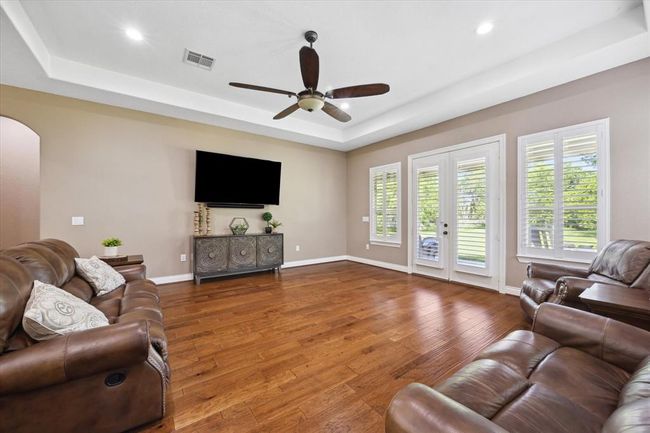 This spacious living room features hardwood floors, neutral walls, and ample natural light from large windows and French doors. It includes a ceiling fan, and comfortable leather seating, offering a welcoming and stylish atmosphere. | Image 14