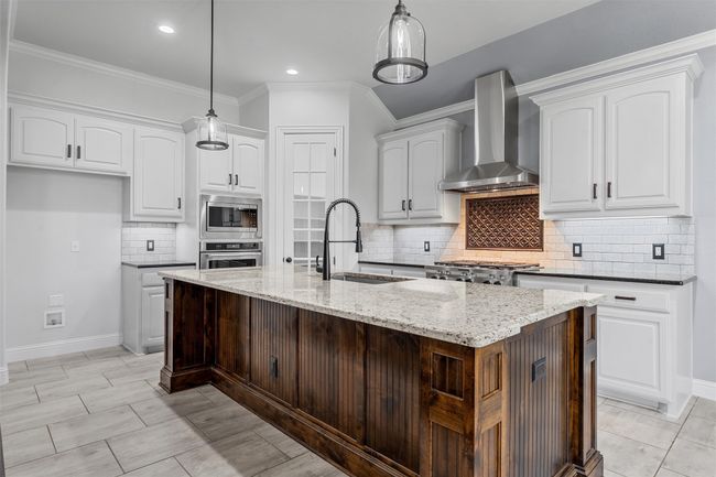 Kitchen featuring wall chimney exhaust hood, stainless steel appliances, ornamental molding, light stone countertops, and white cabinetry | Image 10