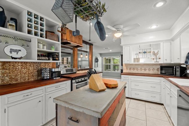 This kitchen features a rustic charm with white cabinetry, wooden countertops, and a central island with a butcher block top. It includes modern appliances, a ceiling fan, and open shelving for storage. The space is well-lit with recessed lighting and has a decorative backsplash for added character. | Image 9